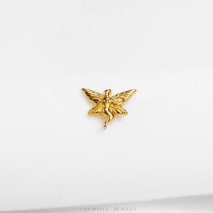 May include: A gold-coloured fairy pendant with outstretched wings. The fairy is depicted in a graceful pose, with delicate details. The pendant is set against a white background, highlighting its intricate design. The jewellery piece is likely a charm or a small pendant.