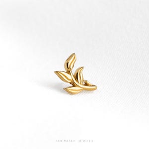 May include: A gold-toned stud earring shaped like a small leafy branch with three distinct leaves. The piece features a polished metallic finish and is positioned against a plain white background. The text SHEMISLI JEWELS is printed at the bottom.