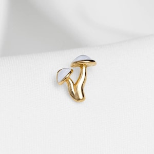 Peut inclure: Un charm en forme de champignon doré avec deux chapeaux. Les chapeaux sont blancs et les tiges sont dorées. Le charm est probablement un pendentif ou une boucle d'oreille, sur fond blanc.