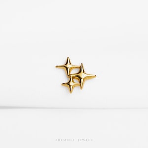 May include: A gold-toned metal stud earring featuring a cluster of three four-pointed star shapes of varying sizes. The surface is smooth and reflective, catching the light. The text "SHEMISLI JEWELS" is printed in small, dark letters at the bottom.