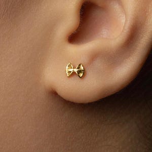 May include: A small, gold-coloured bow-shaped earring is shown on an ear. The earring is a stud style, with a detailed bow design. The earring is a bright, shiny gold colour.