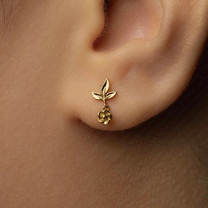 May include: A gold earring with a floral design. The earring features a leaf-shaped top and a small flower pendant. The earring is displayed on an ear, highlighting its delicate details and elegant style.