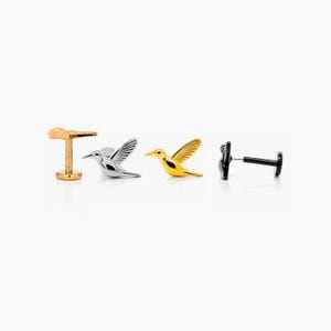 May include: A selection of hummingbird-shaped earrings in gold, silver, and black. The earrings are presented against a white backdrop. The earrings are small and detailed, with the hummingbird's wings spread. Likely made of metal, these earrings are a jewellery item.