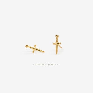 May include: A pair of gold-coloured dagger earrings. Each earring features a detailed handle and a sharp blade. The earrings are set against a white backdrop. The text "SHEMISLI JEWELS" is visible at the bottom of the image.