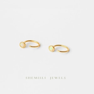May include: A pair of gold-coloured hoop earrings, each featuring a small, round, opalescent stone. The earrings are set against a white backdrop. The text "SHEMISLI JEWELS" is visible at the bottom of the image.