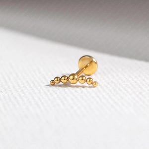 May include: A gold earring featuring a curved line of small, spherical beads. The earring has a flat, circular back and a post for piercing. The beads are evenly spaced, creating a delicate and elegant design.
