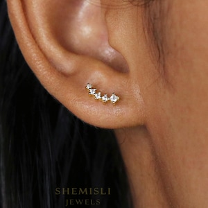 May include: A gold ear climber earring featuring six clear, sparkling stones. The earring curves along the ear, with the stones increasing in size. The brand name "SHEMISLI JEWELS" is visible at the bottom of the image.
