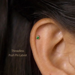 Peut inclure: Gros plan d'une oreille avec une boucle d'oreille labret à tige filetée de couleur or. La boucle d'oreille présente une pierre verte émeraude en forme de goutte d'eau, entourée d'une bordure perlée dorée. Le texte "Threadless Push Pin Labret" est visible.