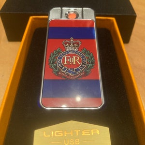 Może przedstawiać: Srebrna zapalniczka USB z czerwono-niebiesko-białym wzorem przedstawiającym herb królewski z tekstem "HONI SOIT QUI MAL Y PENSE" i "ROYAL ENGINEERS" oraz tekstem "LIGHTER - USB" na złotej etykiecie.