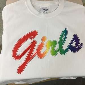 May include: A white t-shirt with the word "Girls" in a rainbow gradient design. The text is in a cursive font, with red at the beginning and transitioning through orange, yellow, green, and blue to purple at the end.