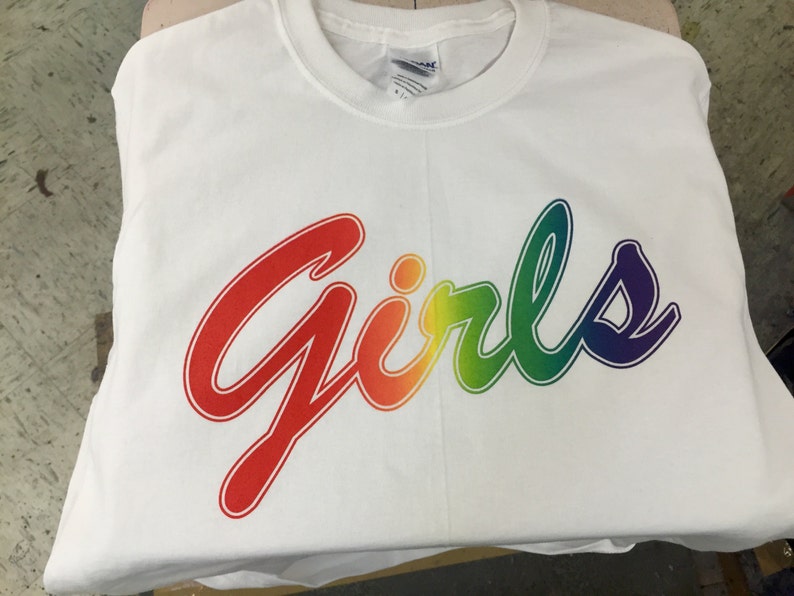 May include: A white t-shirt with the word "Girls" in a rainbow gradient design. The text is in a cursive font, with red at the beginning and transitioning through orange, yellow, green, and blue to purple at the end.
