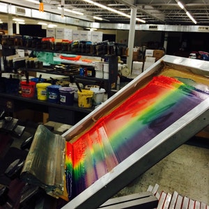May include: A screen printing setup with a rainbow of colors being applied to a surface. The image shows a screen printing frame with a squeegee, and various paint buckets in the background. The colors include red, orange, yellow, green, blue, and purple.