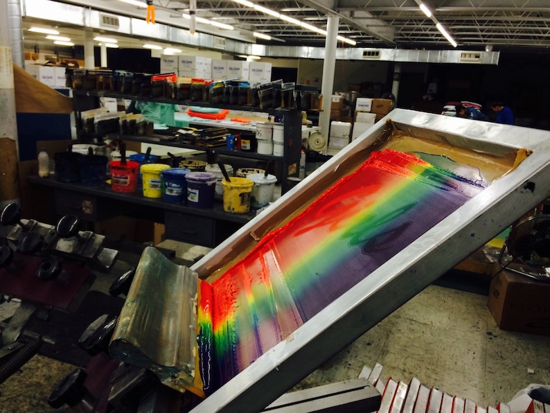 May include: A screen printing setup with a rainbow of colors being applied to a surface. The image shows a screen printing frame with a squeegee, and various paint buckets in the background. The colors include red, orange, yellow, green, blue, and purple.