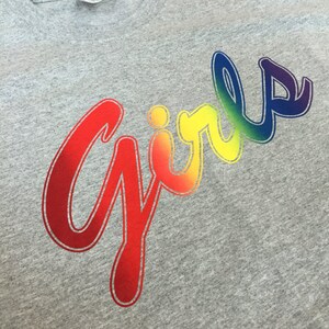 May include: A heather gray t-shirt with the word "Girls" in a rainbow gradient design. The word is written in a cursive font, with the colors transitioning from red to purple. The shirt has a crew neck and short sleeves.