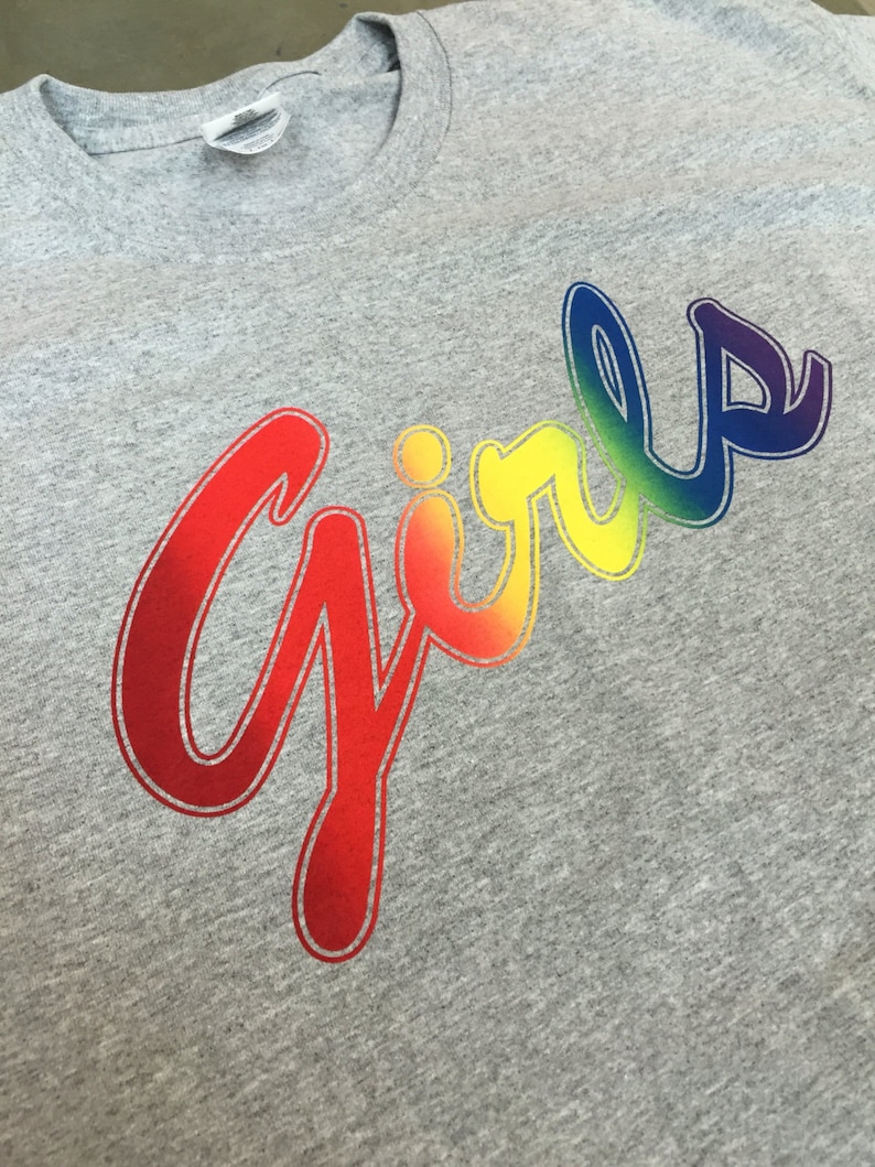 May include: A heather gray t-shirt with the word "Girls" in a rainbow gradient design. The word is written in a cursive font, with the colors transitioning from red to purple. The shirt has a crew neck and short sleeves.