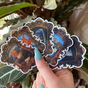 May include: A selection of stickers depicting animal parodies of famous paintings. Each sticker presents a different animal portrait within a decorative, brown frame. The stickers are held in a hand with blue nail polish, set against green foliage.