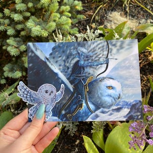 May include: A fantasy art print featuring a figure riding a snowy owl with a bow and arrow, set against a snowy mountain backdrop. A small owl sticker is held in front of the print. The artwork is predominantly blue and white.