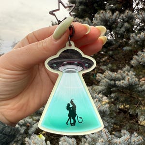 May include: A keychain featuring a UFO design with a star-shaped clasp. The UFO emits a green beam of light, illuminating a silhouette of a couple. The keychain is a unique accessory.
