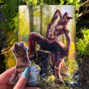 May include: A print featuring a mythological satyr figure with horns, seated on a tree stump in a forest setting. A sticker of a similar character is held in front of the print. The artwork has a fantasy theme with vibrant colors and detailed illustrations.