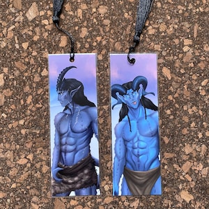 May include: Two rectangular bookmarks featuring illustrations of blue-skinned fantasy figures with horns and long black hair. Each bookmark has a black tassel attached at the top. The figures are depicted in a muscular physique, wearing loincloths, against a purple sky.