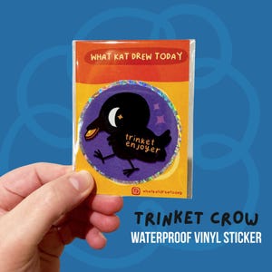May include: A waterproof vinyl sticker with a black crow design and the words "trinket enjoyer" on a purple circle. The sticker is on a yellow and orange card with the text "WHAT KAT DREW TODAY".