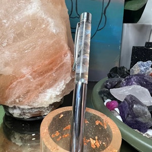 May include: A silver pen with a clear body filled with white and gray swirls. The pen is standing upright in a clear glass holder with gold flecks.