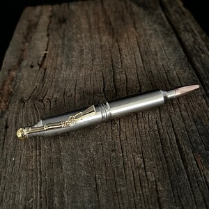 May include: A silver pen with gold accents, designed to resemble a bullet. The pen features a copper-colored tip and intricate gold detailing near the pen's tip. The pen is set against a rustic, weathered wooden surface.