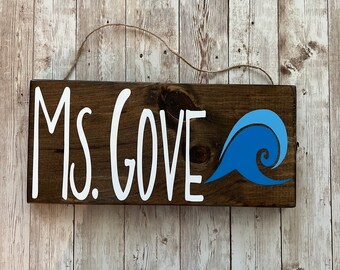May include: A dark brown wooden sign with white lettering that reads "Ms. Gove" and a blue wave graphic.