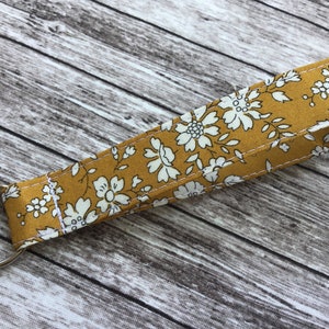 Puede incluir: Un llavero de tela amarillo mostaza con un patr&oacute;n floral blanco. El llavero tiene un anillo de metal plateado.