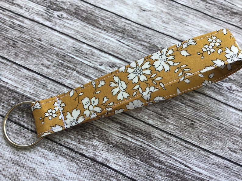 Puede incluir: Un llavero de tela amarillo mostaza con un patr&oacute;n floral blanco. El llavero tiene un anillo de metal plateado.