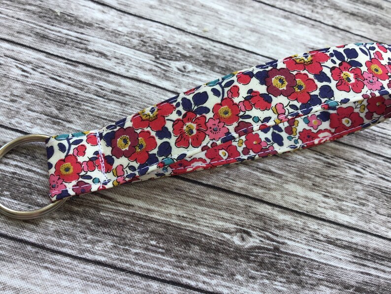 Puede incluir: Un llavero de tela blanca con un patr&oacute;n floral en tonos rojos, rosas, azules y amarillos. El llavero tiene un anillo de metal plateado.