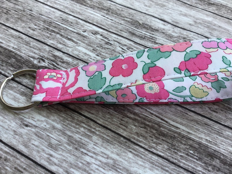 Puede incluir: Un llavero de tela blanca con un estampado floral rosa. El llavero tiene un anillo de metal plateado.