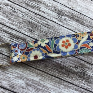 Puede incluir: Un llavero con estampado floral colorido con un anillo de llave plateado. La tela es una mezcla de azul, naranja, amarillo y blanco.