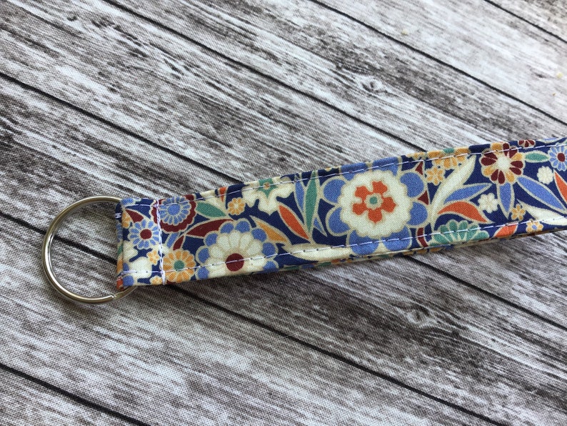 Puede incluir: Un llavero con estampado floral colorido con un anillo de llave plateado. La tela es una mezcla de azul, naranja, amarillo y blanco.
