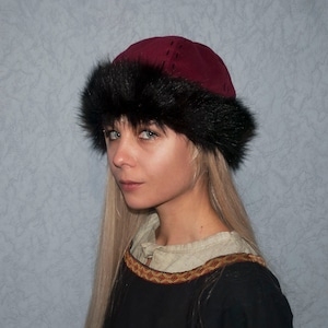 May include: A woman wearing a burgundy suede hat with a black fur trim. The hat has a stitched design on the top. She is wearing a black tunic with a gold and red trim.