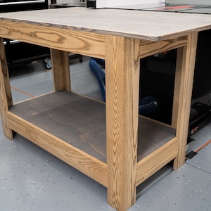 May include: A wooden workbench with a light brown stain and a gray top. The workbench has a lower shelf and is made of solid wood.