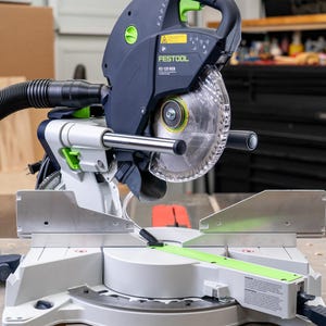 May include: A Festool TS 55 REQ saw with a dust collection hose attached. The saw is on a table with a green and gray cutting guide. The saw has a clear plastic blade guard.