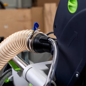 May include: A close-up of a clear plastic dust collection hose connected to a black and green power tool. The hose is attached to the tool with a blue clamp.