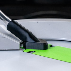 May include: Close-up of a black plastic dust collection port on a table saw. The port is connected to a flexible hose and is positioned over a green cutting guide.