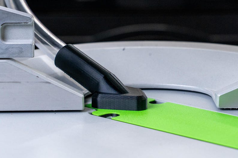 May include: Close-up of a black plastic dust collection port on a table saw. The port is connected to a flexible hose and is positioned over a green cutting guide.