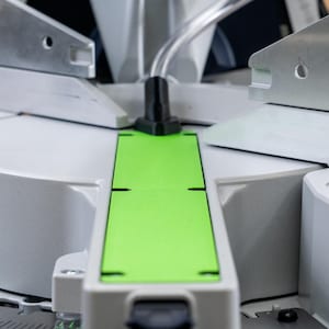 May include: Close-up of a white and gray machine with a green strip. The machine has a circular base and a series of metal arms.