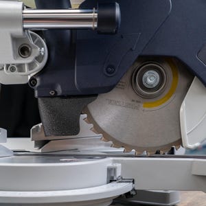 May include: Close-up of a silver circular saw blade with sharp teeth, mounted on a dark blue and gray miter saw. The blade has yellow and black markings and the brand name "FESTOOL" is visible. The saw is designed for precision cutting of wood and other materials.