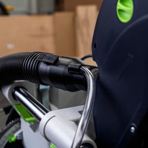May include: Close-up of a black dust collection hose connected to a Festool KS 120 saw. The hose has a black ribbed texture and a green and black connector.