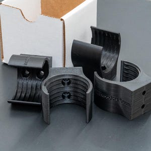May include: Four black plastic pipe clamps with a ribbed texture. The clamps are designed to hold pipes or other round objects securely in place.