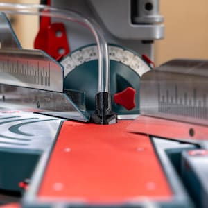 May include: Close-up of a miter saw with a red base and silver blade guides. The saw features a clear plastic tube and a black fitting. A degree scale is visible on the teal-colored base, with a red lever.
