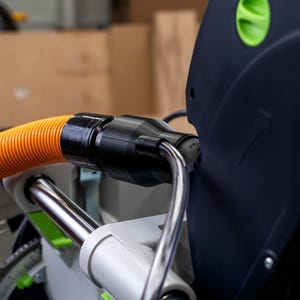 May include: Close-up of a black and orange dust collection hose connected to a Festool KS 120 saw. The hose is connected to the saw with a black connector.