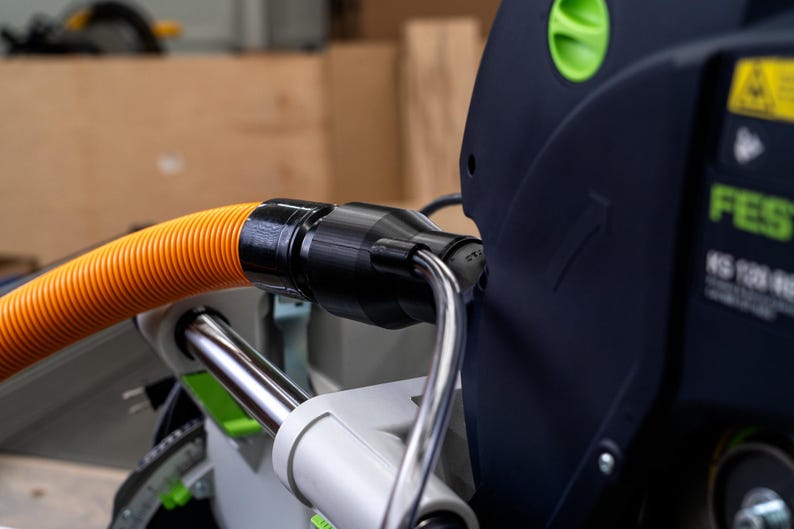 May include: Close-up of a black and orange dust collection hose connected to a Festool KS 120 saw. The hose is connected to the saw with a black connector.