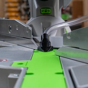 Op de afbeelding: Close-up van een grijze en groene verstekzaag. De zaag heeft een zwart kunststof onderdeel en een doorzichtige buis. De basis is felgroen. De zaag is ontworpen voor het zagen van hout en andere materialen.