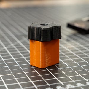 May include: A 3D-printed orange and black object. The object has a rectangular orange base with a black, flower-shaped top. The top has a hole in the center. The object is sitting on a gray surface with a grid pattern.