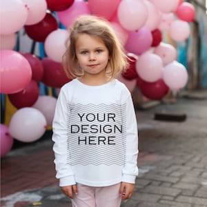 May include: A young child wearing a white long-sleeved shirt with the text "YOUR DESIGN HERE" in black. The shirt has a black zigzag design. The background features pink and maroon balloons.